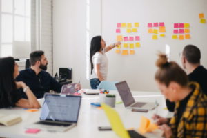 Business meeting with 4 people around a table one laptop opened. One woman is standing by a whiteboard learning the mistakes from a family business succession meeting